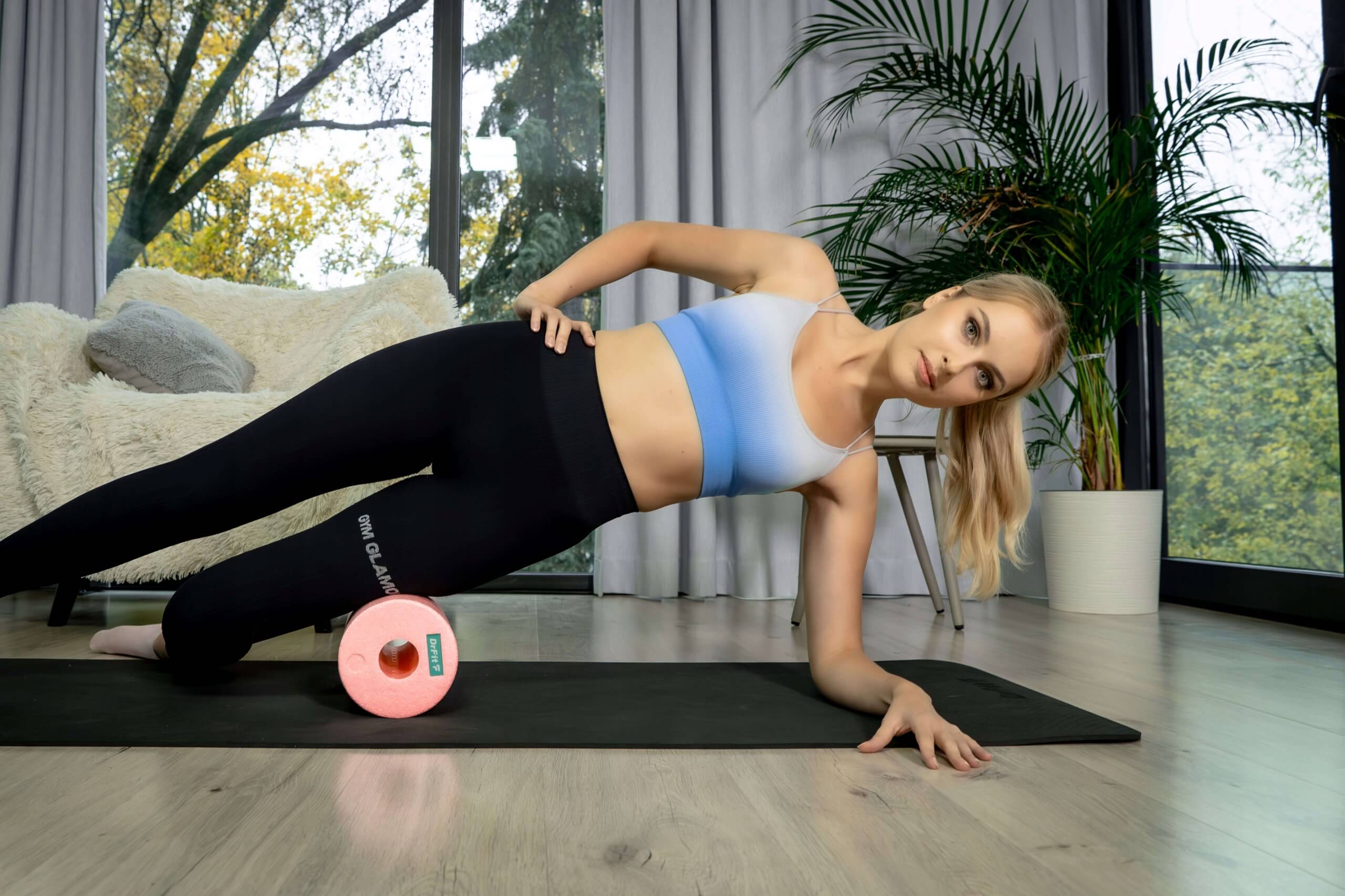 Kobieta wykonująca boczne rolowanie uda na różowym rollerze DrFit, w pozycji podpartego boku na macie treningowej w domowym wnętrzu.